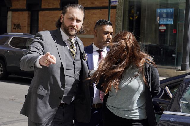 Lauren Pazienza, whose red hair is covering her face, has her hands behind her back with two detectives in suits guiding her
